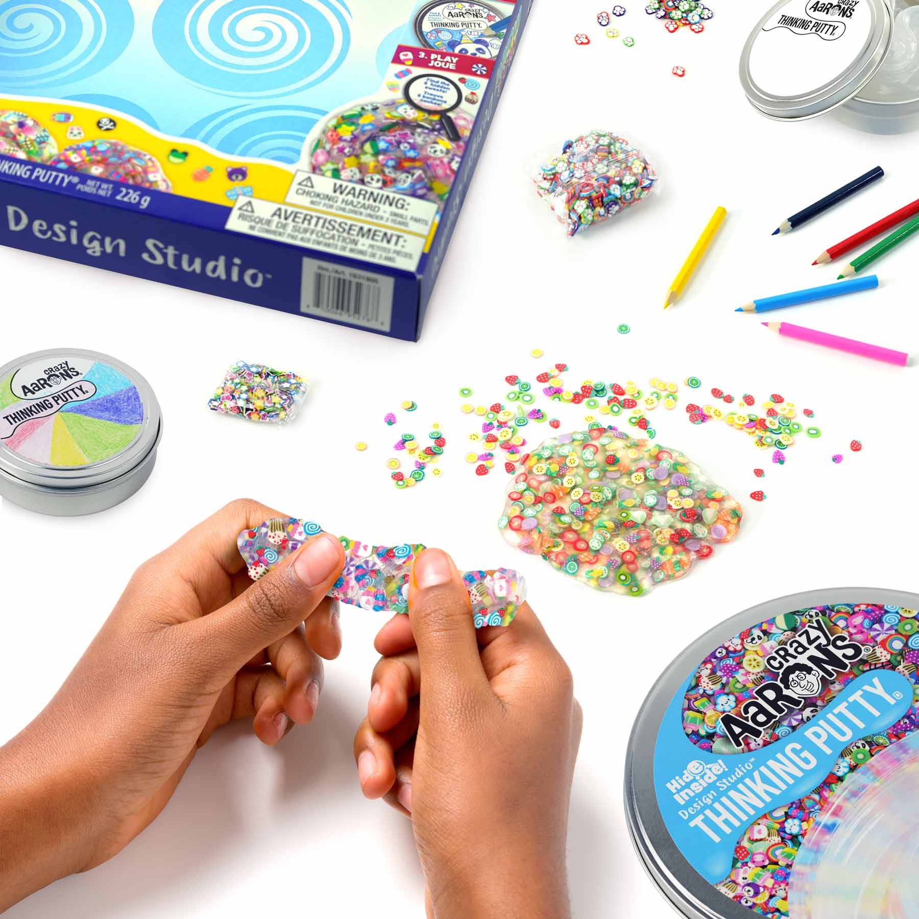 Colorful Thinking Putty is being stretched by hands on a table surrounded by craft supplies, putty containers, and small beads. A box labeled "Crazy Aarons Hide Inside Design Studio Kit" from Crazy Aarons, along with colorful pens, sets up the perfect scene for sensory integration fun.