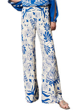 The person is elegantly dressed in the Figue Devon Floral Pant, featuring a blue and white floral pattern with a signature straight silhouette and convenient elastic waistband, paired with a matching blue top.