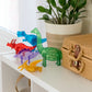 The MAGNA-TILES® Dinos 5-Piece Set by Magnatiles, featuring five colorful translucent dinosaur figurines compatible with the MAGNA-TILES® system, is displayed on a white shelf beside a potted plant and a woven basket.