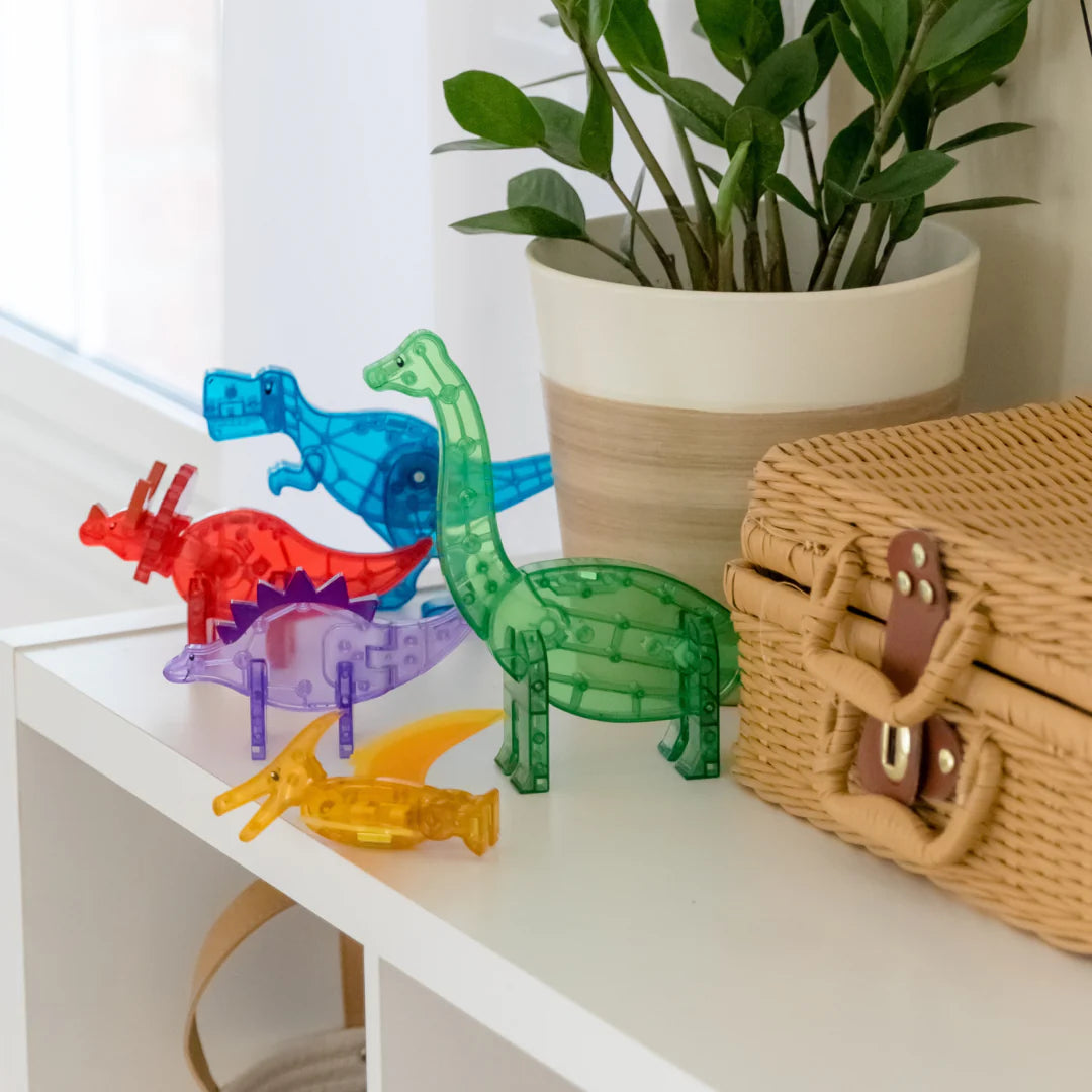 The MAGNA-TILES® Dinos 5-Piece Set by Magnatiles, featuring five colorful translucent dinosaur figurines compatible with the MAGNA-TILES® system, is displayed on a white shelf beside a potted plant and a woven basket.