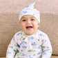A smiling baby wearing the Kissy Kissy Dinosaur Delight Hat sits on a light wood floor in front of a beige textured bed, dressed in dinosaur-patterned Pima cotton pajamas.