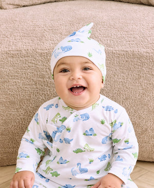 A baby smiles while sitting on a beige floor in front of a textured beige couch, wearing cozy white pajamas and the Kissy Kissy “Baby Dinosaur Delight Hat” featuring playful blue and green dinosaur prints.