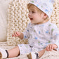 A toddler in the Kissy Kissy Baby Boys’ Dinosaur Era Playsuit, featuring blue and green patterns, with a matching hat, sits smiling on a knitted blanket and looks to the side.