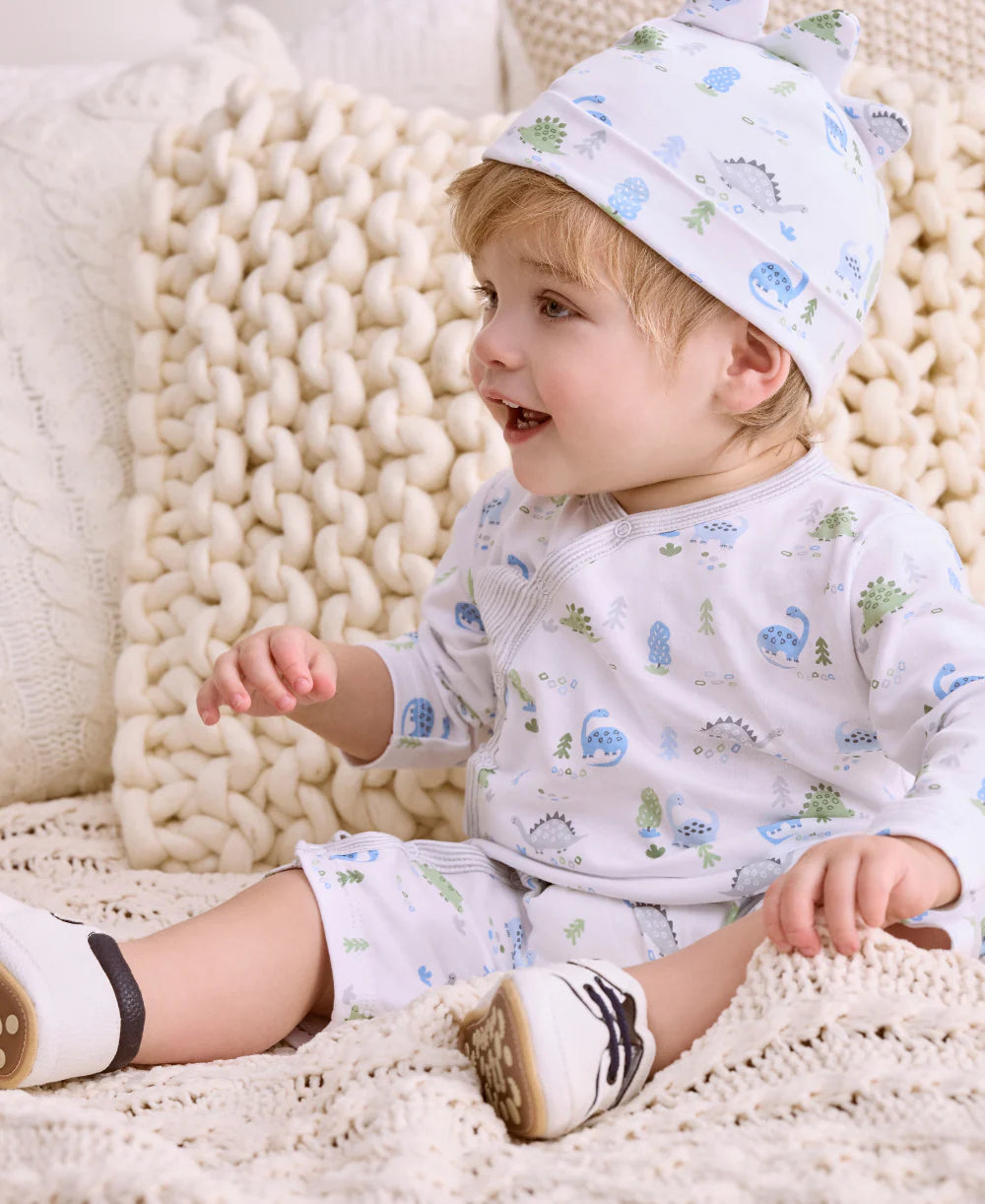 A toddler in the Kissy Kissy Baby Boys’ Dinosaur Era Playsuit, featuring blue and green patterns, with a matching hat, sits smiling on a knitted blanket and looks to the side.