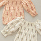 Two Noralee Baby Girls' Autumn Rompers with smocked empire bodice and bubbled long sleeves, one peach with red flowers, the other white with grey flowers, are laid flat on a textured beige surface.