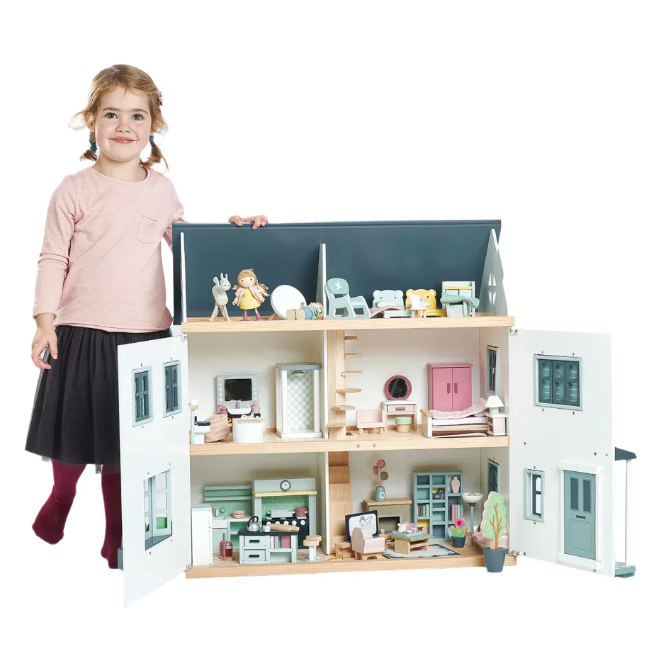 A child stands beside the open Tender Leaf Dovetail Doll House by Tender Leaf Toys, its rooms elegantly furnished with dovetail pieces in gender-neutral hues.