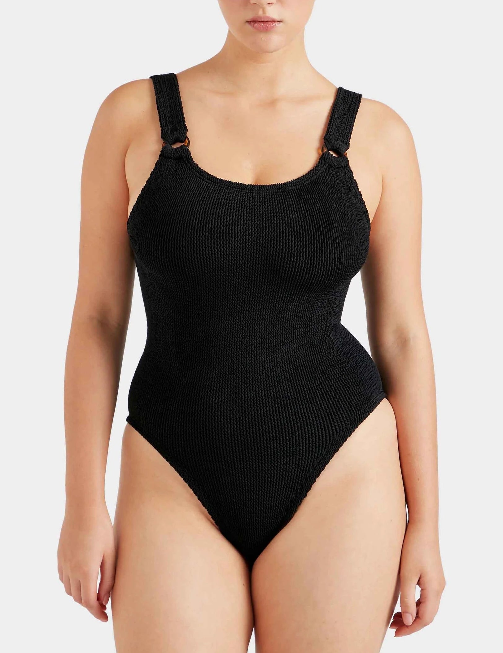 A woman wears the Hunza G Domino Swim, a black crinkle one-piece with wide straps and high-cut legs, shown from the front against a plain background.