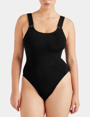A woman wears the Hunza G Domino Swim, a black crinkle one-piece with wide straps and high-cut legs, shown from the front against a plain background.