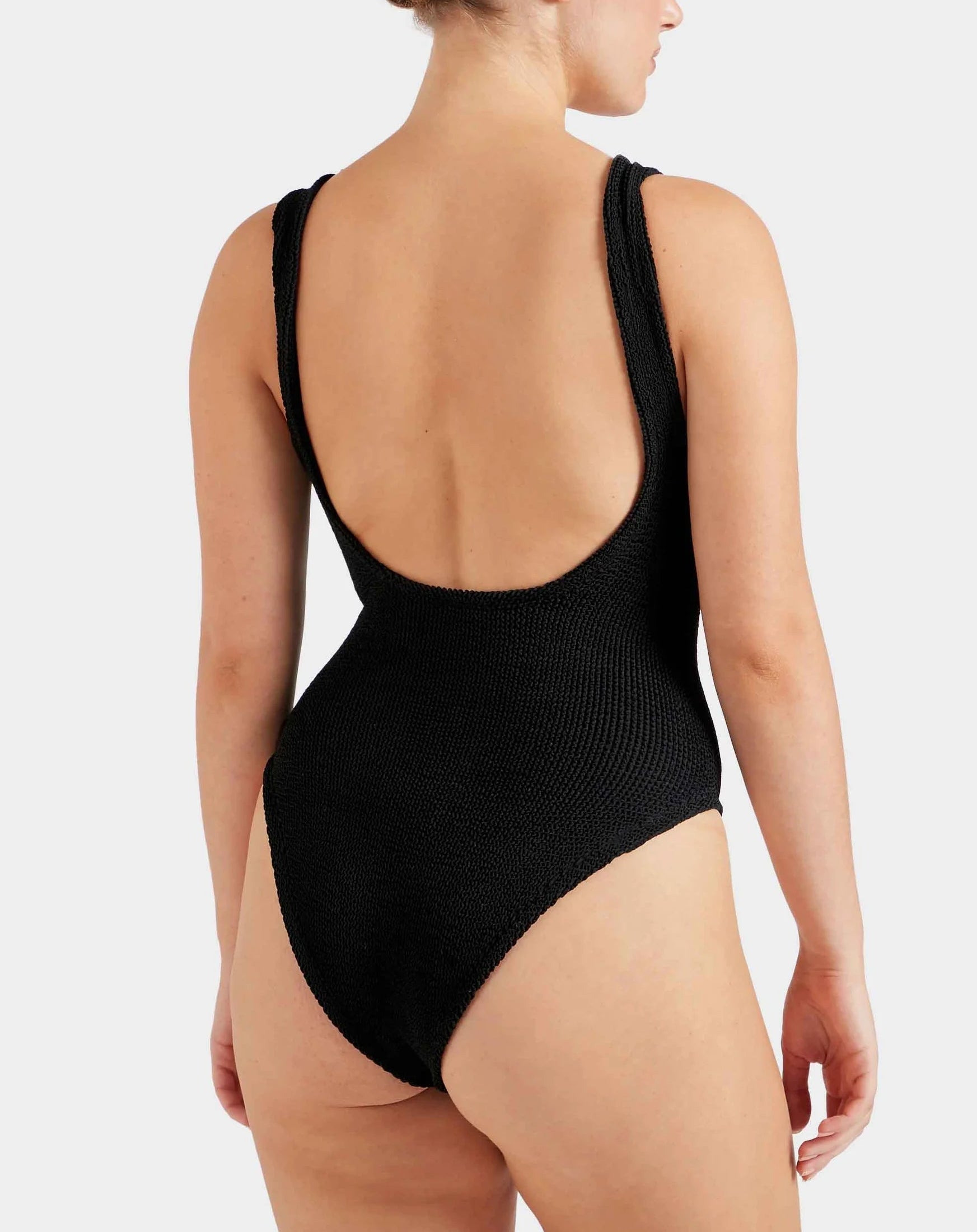 A person is wearing the Hunza G Domino Swim, a black crinkle-textured one-piece swimsuit with a low scoop back, shown from behind against a plain background.