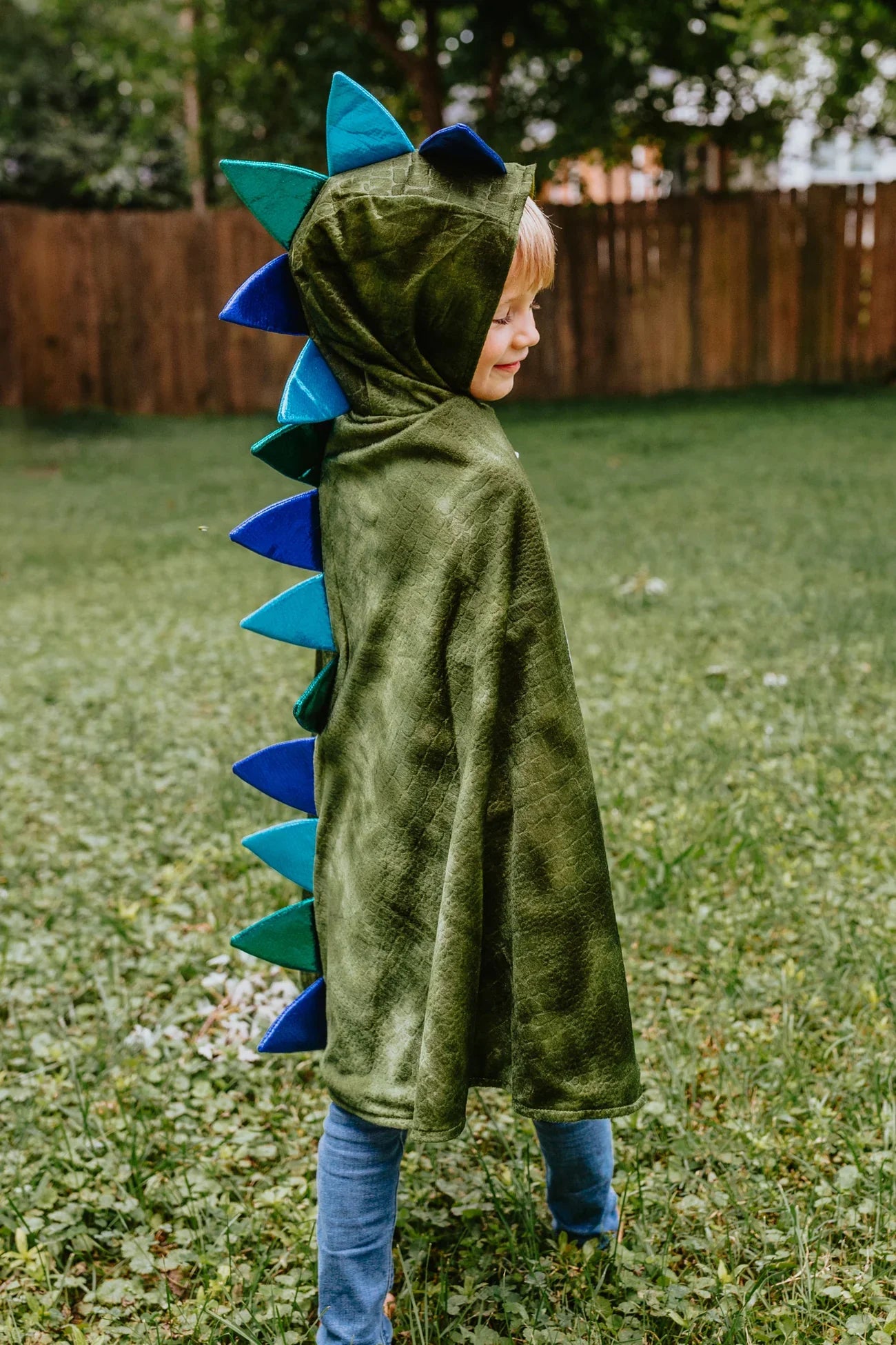 A child wearing the Great Pretenders Dragon Cape with Claws in Green, adorned with blue spikes and made of crushed velvet, stands on the grass in the backyard.