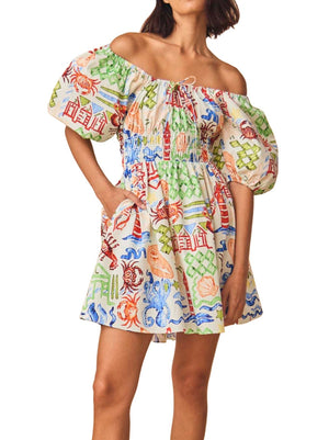 A woman wearing a Hunter Bell NYC Drew Dress, a colorful off-shoulder mini dress featuring a vibrant print with various motifs and a scoop neckline.