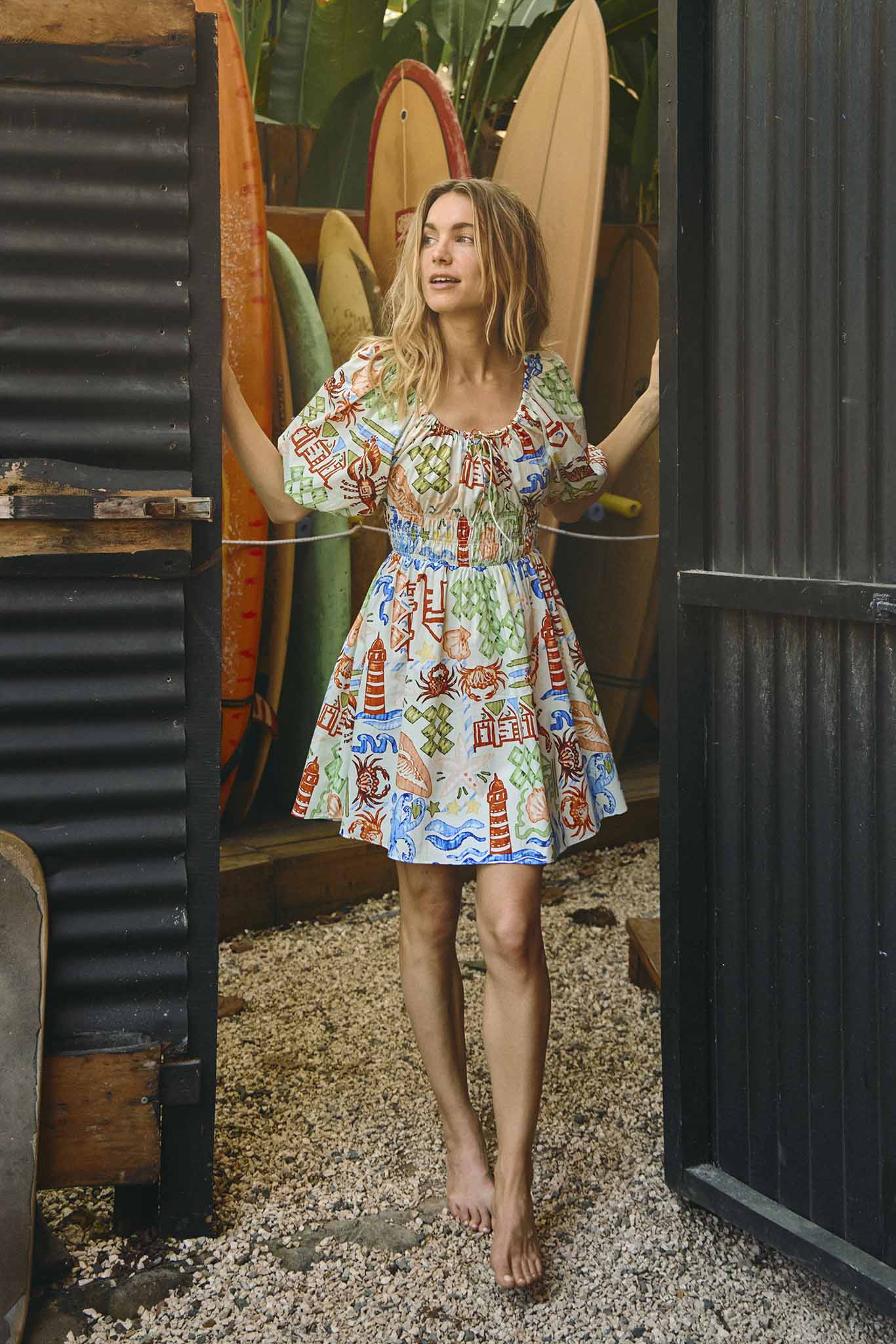 A woman in a Hunter Bell NYC Drew dress with a smocked elastic waist stands barefoot between surfboards, exiting a dark container into a lush garden.