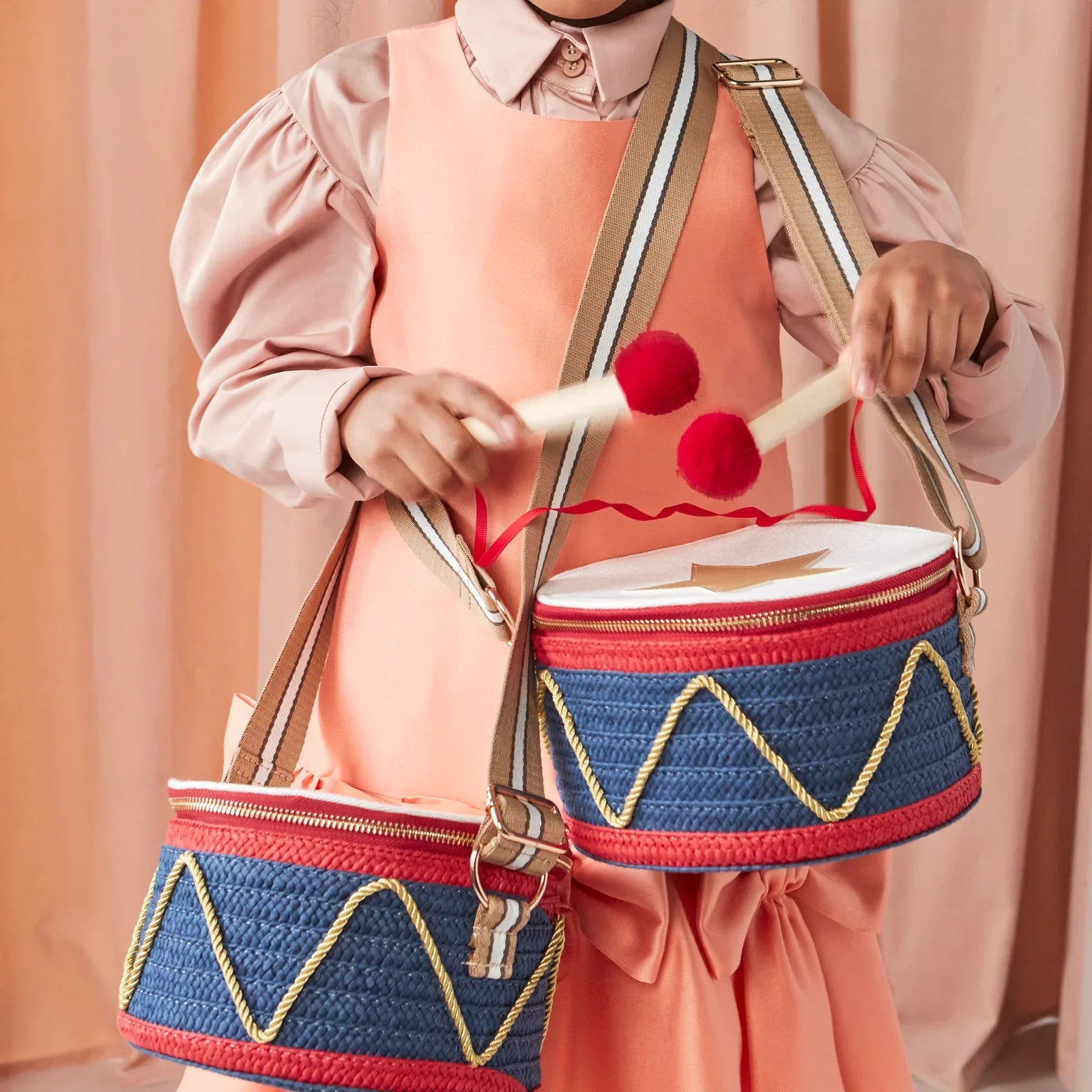 A child dressed in a peach outfit is playing with the Meri Meri Drum Bag, featuring two blue and red toy drums and wooden drumsticks.