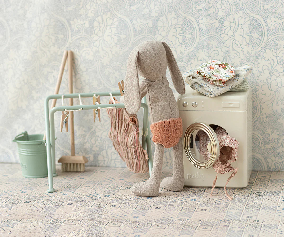 A stuffed bunny stands in front of a toy washing machine with a piece of fabric hanging out, mimicking an adorable laundry routine. In the background, there is a Maileg Drying Rack with Pegs, Miniature by Maileg with hanging clothes, a bucket, and a brush, creating the perfect scene for teddies and bunnies.