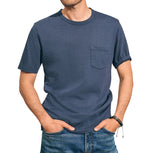 A person wearing a Faherty Sunwashed Pocket Tee from Faherty crafted from organic cotton, complete with a front pocket, and blue jeans stands with hands in their pockets against a neutral background, showcasing a relaxed silhouette.