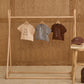Three Rylee & Cru Baby Girls' and Girls' Vintage Tees in beige, gray, and brown with raw edges hang on a wooden rack; a matching brown cap sits on a stool against a wood-paneled wall.