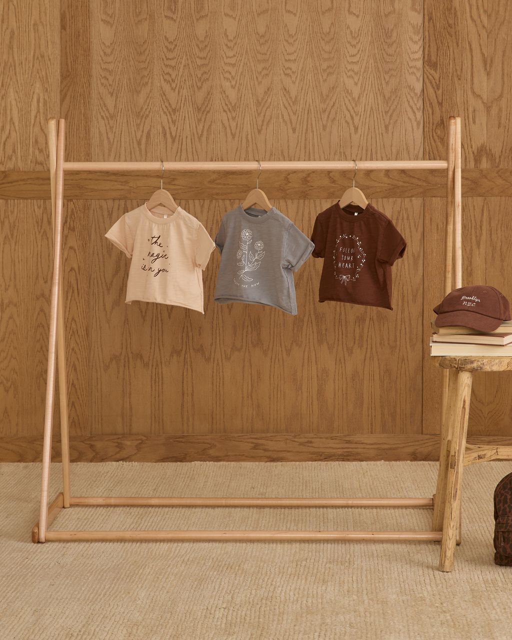 Three Rylee & Cru Baby Girls' and Girls' Vintage Tees in beige, gray, and brown with raw edges hang on a wooden rack; a matching brown cap sits on a stool against a wood-paneled wall.