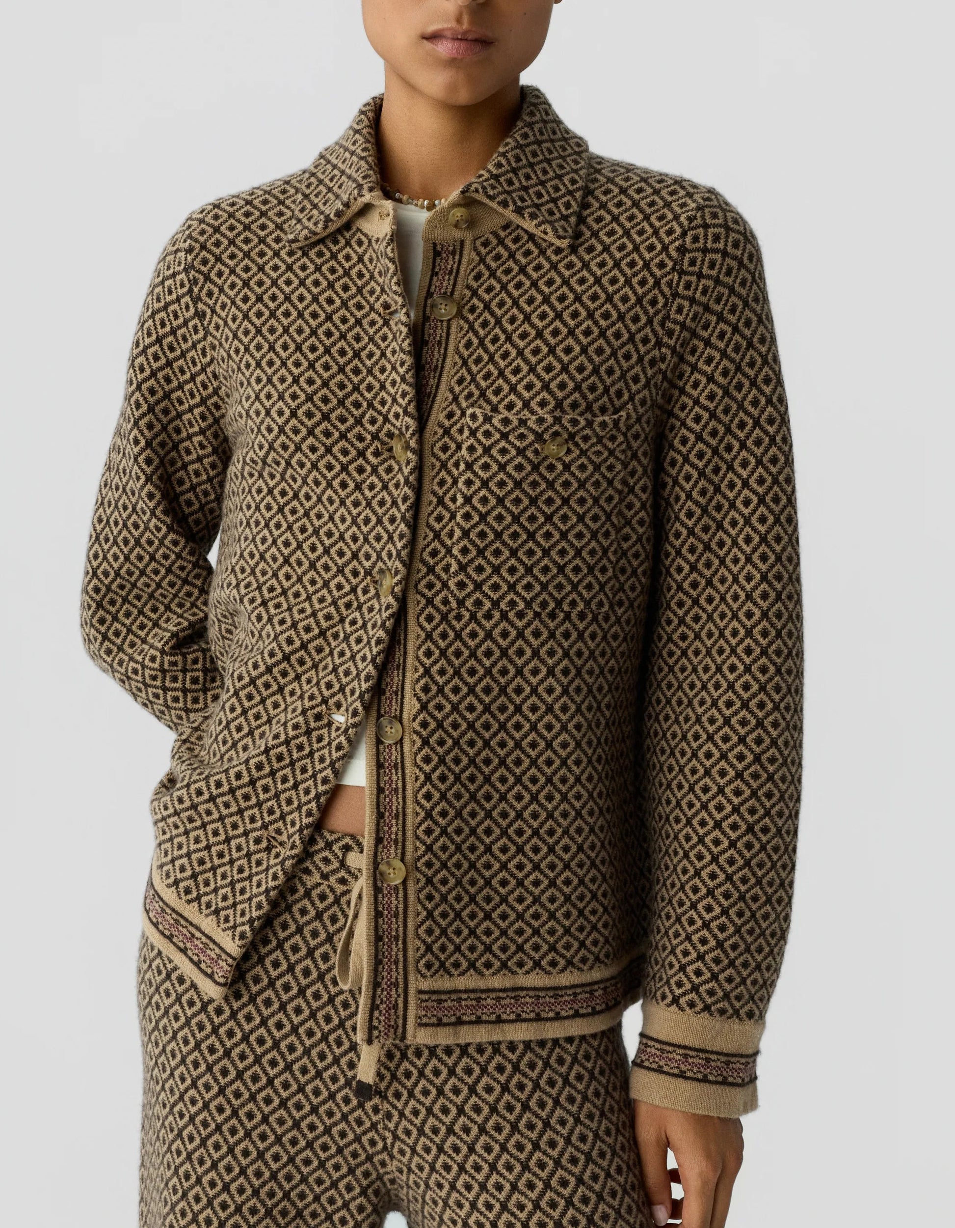 A person wears the Dylan Marquise Shirt by Guest in Residence—a relaxed fit cashmere shirt—layered under a brown and beige patterned button-up jacket with matching pants, both featuring a diamond motif and ribbed trim details for added comfort and style.