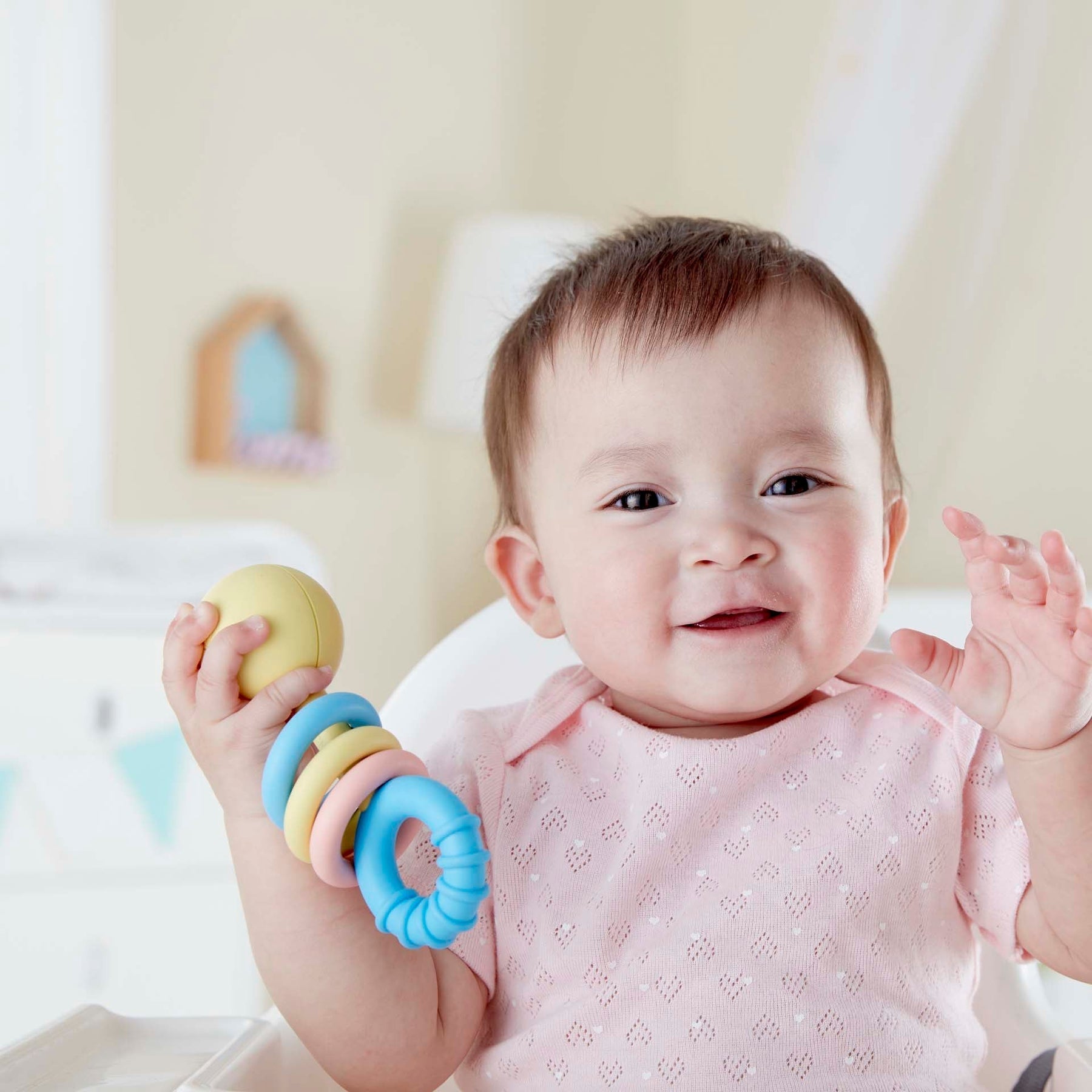 A smiling baby in a pink outfit joyfully holds the colorful, child-safe *Rattling Rings Teether* from Hape while sitting.