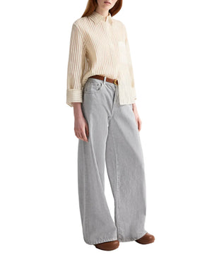 A person wears the Boyfriend Silk Top by TWP in cream, tucked into light gray wide-leg jeans with brown shoes for a relaxed fit.