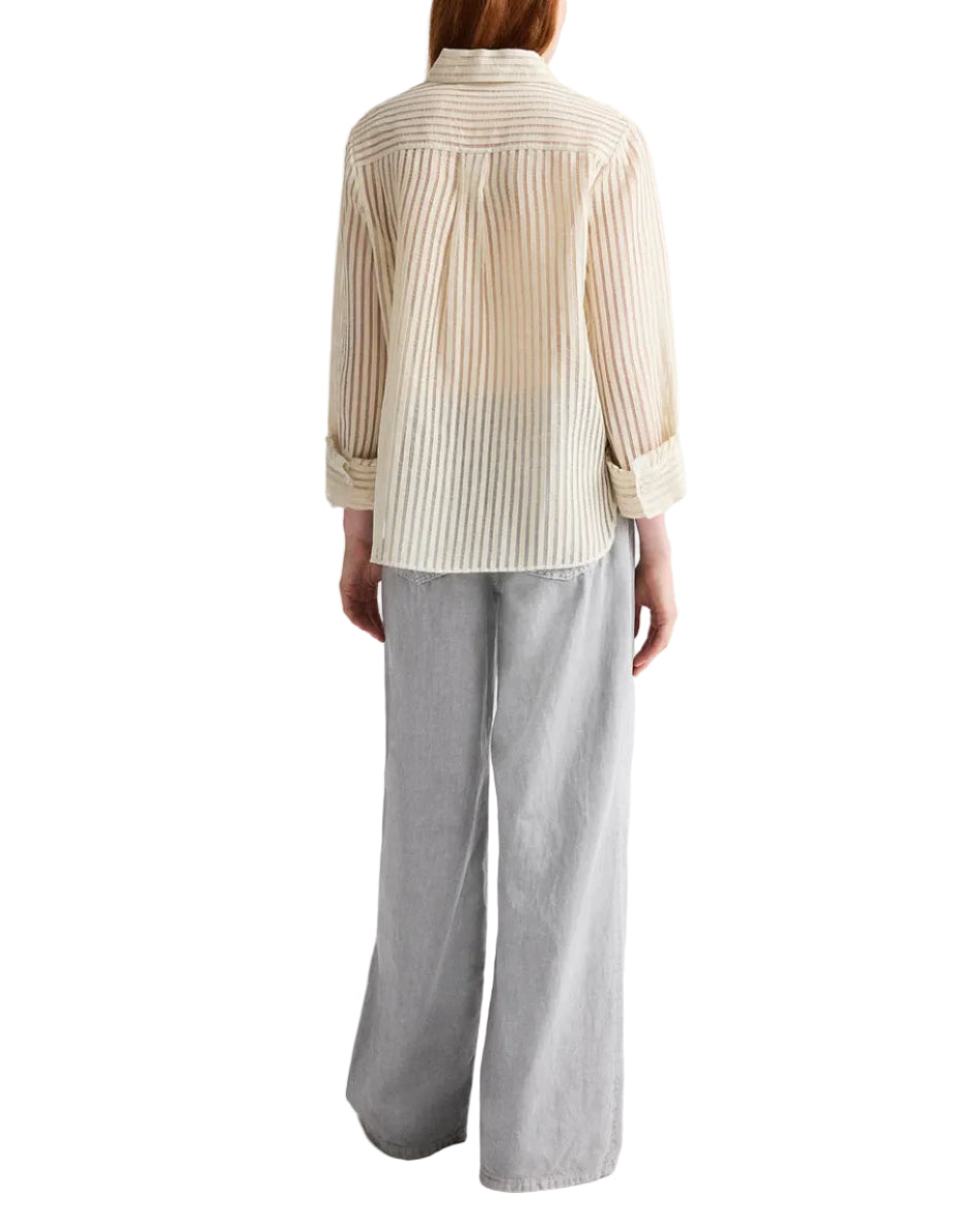 A person stands with their back to the camera, wearing TWP’s Boyfriend Silk Top in beige stripes and loose-fitting light gray pants, set against a white background.