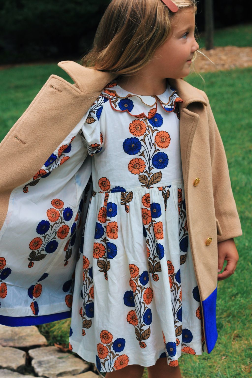 Young girl stands on grass, wearing a tan coat over the Pink Chicken Girls' Darcy Dress with blue and orange patterns, looking to the side.