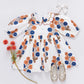 The Pink Chicken Girls' Darcy Dress, featuring a white Buttercup Floral print with orange and blue accents, is styled with a white hair bow, glittery shoes, and red artificial flowers on a white background.