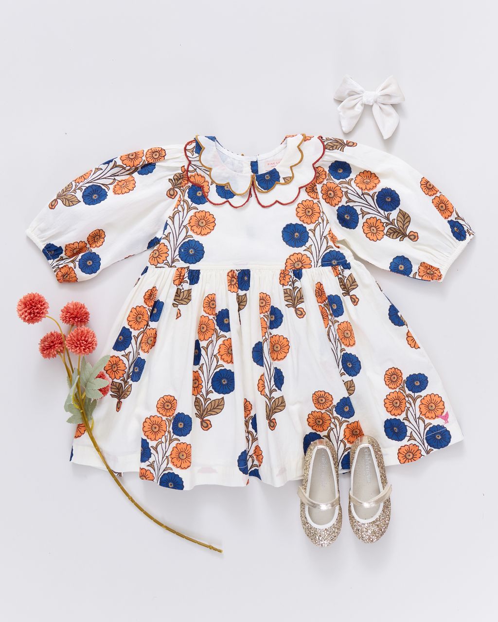 The Pink Chicken Girls' Darcy Dress, featuring a white Buttercup Floral print with orange and blue accents, is styled with a white hair bow, glittery shoes, and red artificial flowers on a white background.