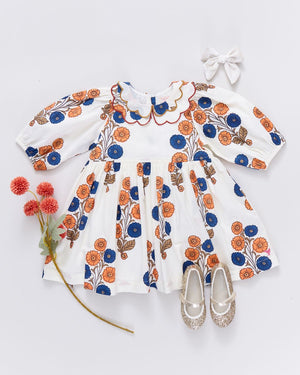 The Pink Chicken Girls' Darcy Dress, featuring a white Buttercup Floral print with orange and blue accents, is styled with a white hair bow, glittery shoes, and red artificial flowers on a white background.