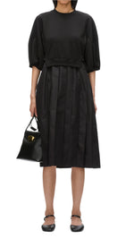 A person is wearing the 3.1 Phillip Lim Lantern Sleeve Sweatshirt Dress, which features lantern sleeves and a pleated skirt, and they are holding a small black handbag. Additionally, they have on black flat shoes with ankle straps.