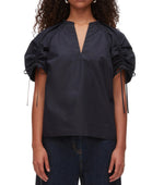 A person wears the 3.1 Phillip Lim Ruched Sleeve V-Neck Top in black, styled with dark jeans and standing against a plain background.