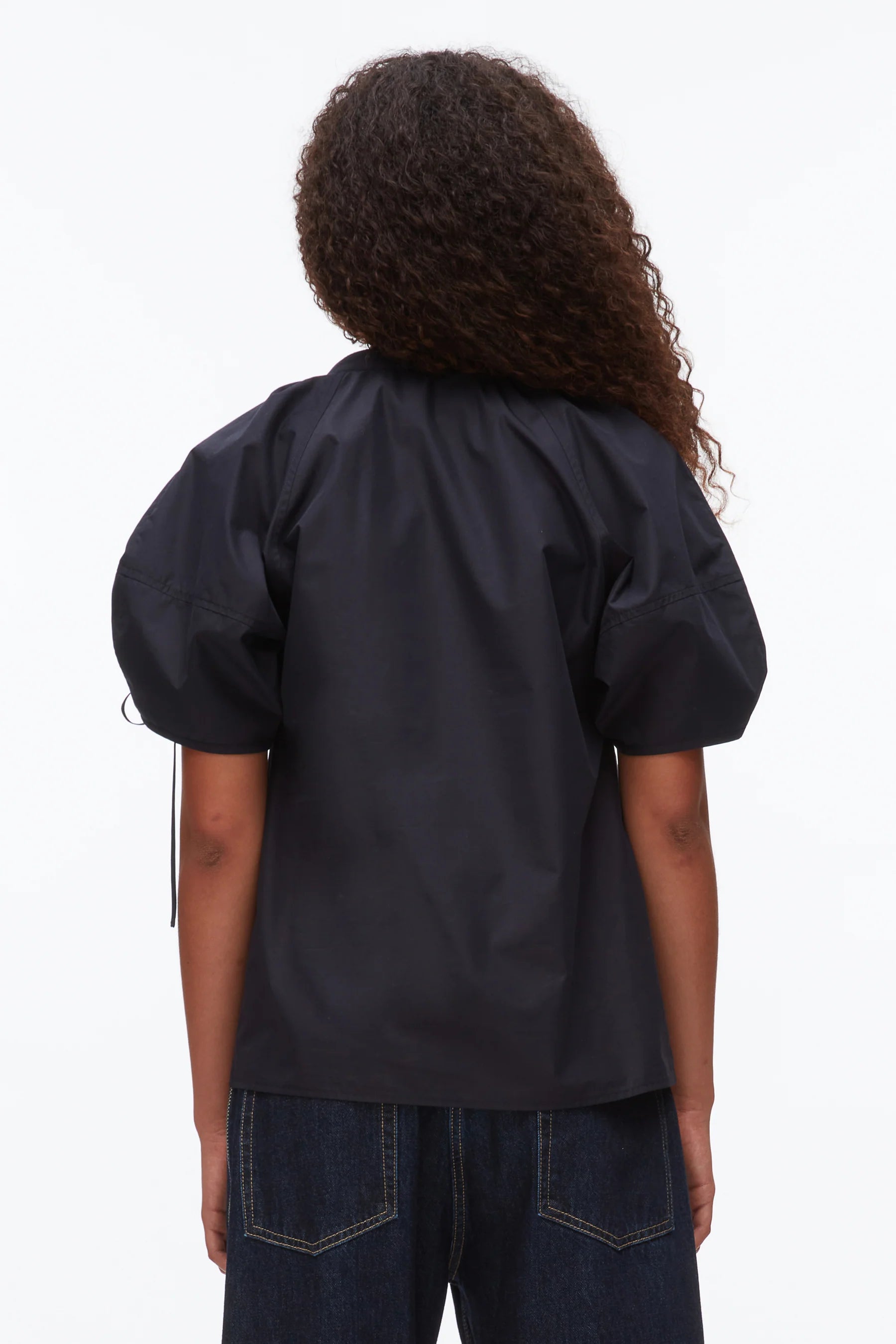 Person with curly hair wears the 3.1 Phillip Lim Ruched Sleeve V-Neck Top in a dark shade with dark denim jeans, shown from the back against a plain background.