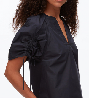 A woman wears the 3.1 Phillip Lim Ruched Sleeve V-Neck Top, a dark short-sleeve blouse in technical poplin with ruched sleeves and a V-neck—combining elegance with modern style. Shown from the chest up.