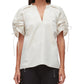 A woman wears the 3.1 Phillip Lim Ruched Sleeve V-Neck Top, a loose white blouse with ruched, drawstring sleeves, paired with black pants featuring metal eyelet details.