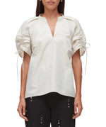 A woman wears the 3.1 Phillip Lim Ruched Sleeve V-Neck Top, a loose white blouse with ruched, drawstring sleeves, paired with black pants featuring metal eyelet details.