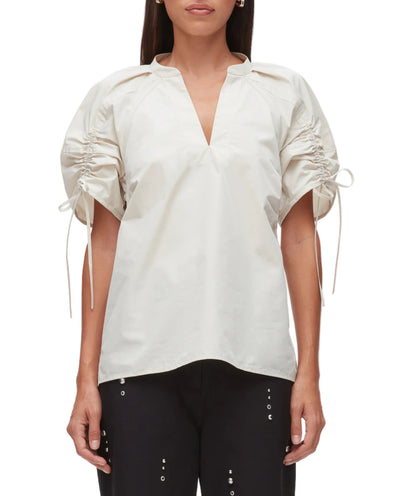 A woman wears the 3.1 Phillip Lim Ruched Sleeve V-Neck Top, a loose white blouse with ruched, drawstring sleeves, paired with black pants featuring metal eyelet details.