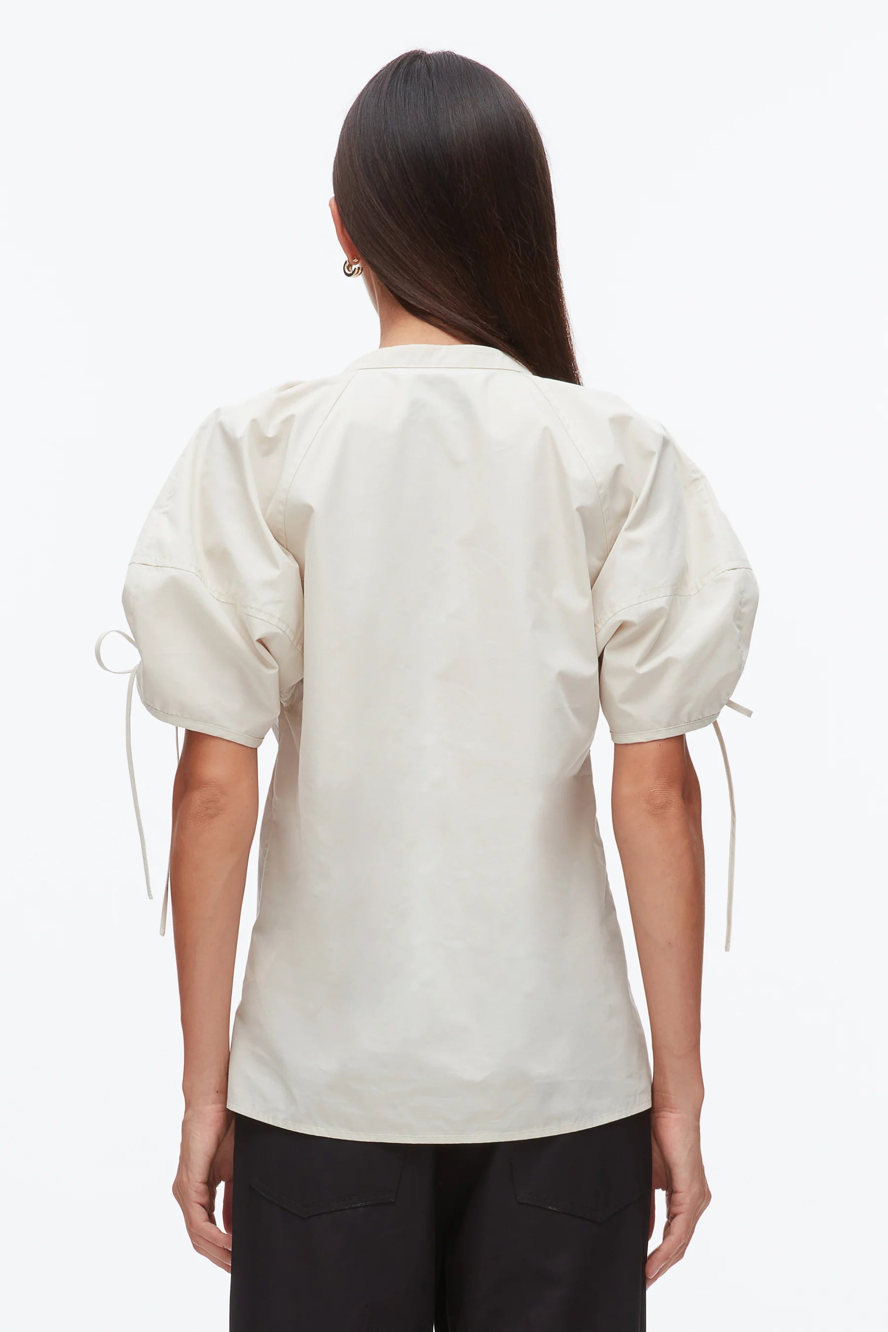 A woman with long dark hair is seen from behind wearing the 3.1 Phillip Lim Ruched Sleeve V-Neck Top in a light hue, paired with black pants.