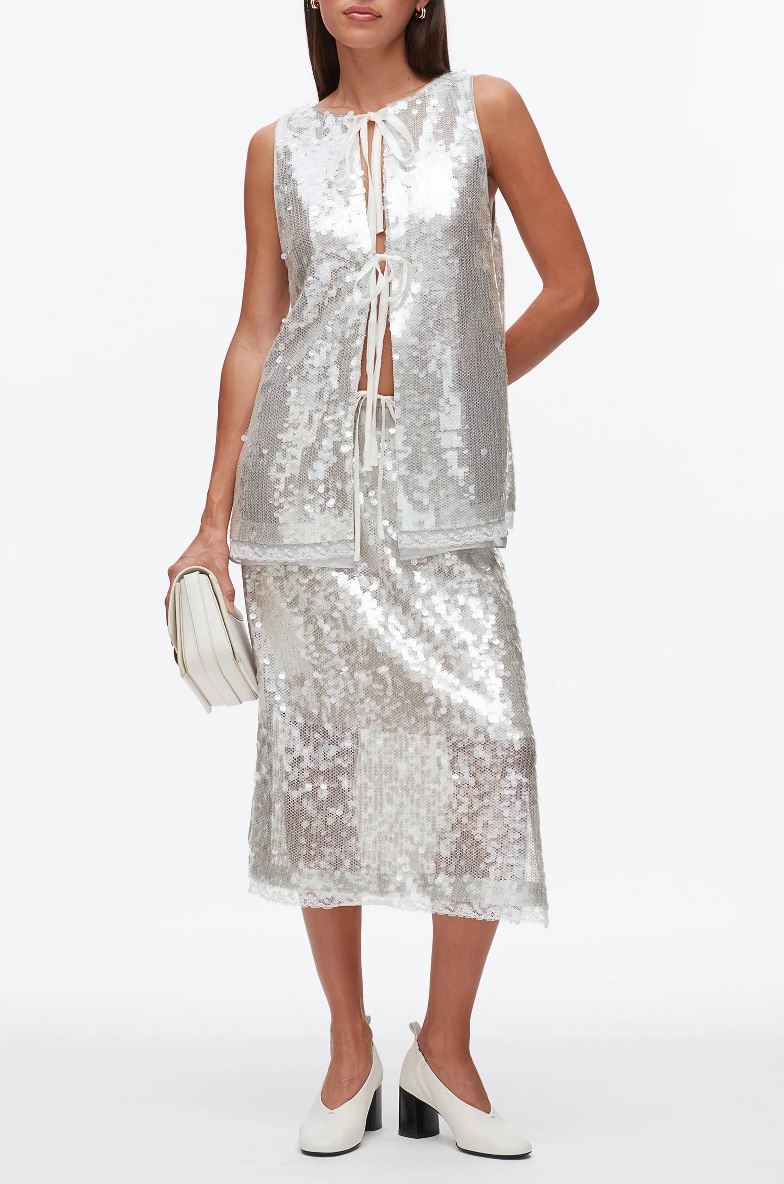 A woman wears the 3.1 Phillip Lim Sequin Flounce Tank with Bows, paired with a matching skirt featuring a lace-trimmed organza hem, and accessorizes with a white clutch and white block-heel shoes.