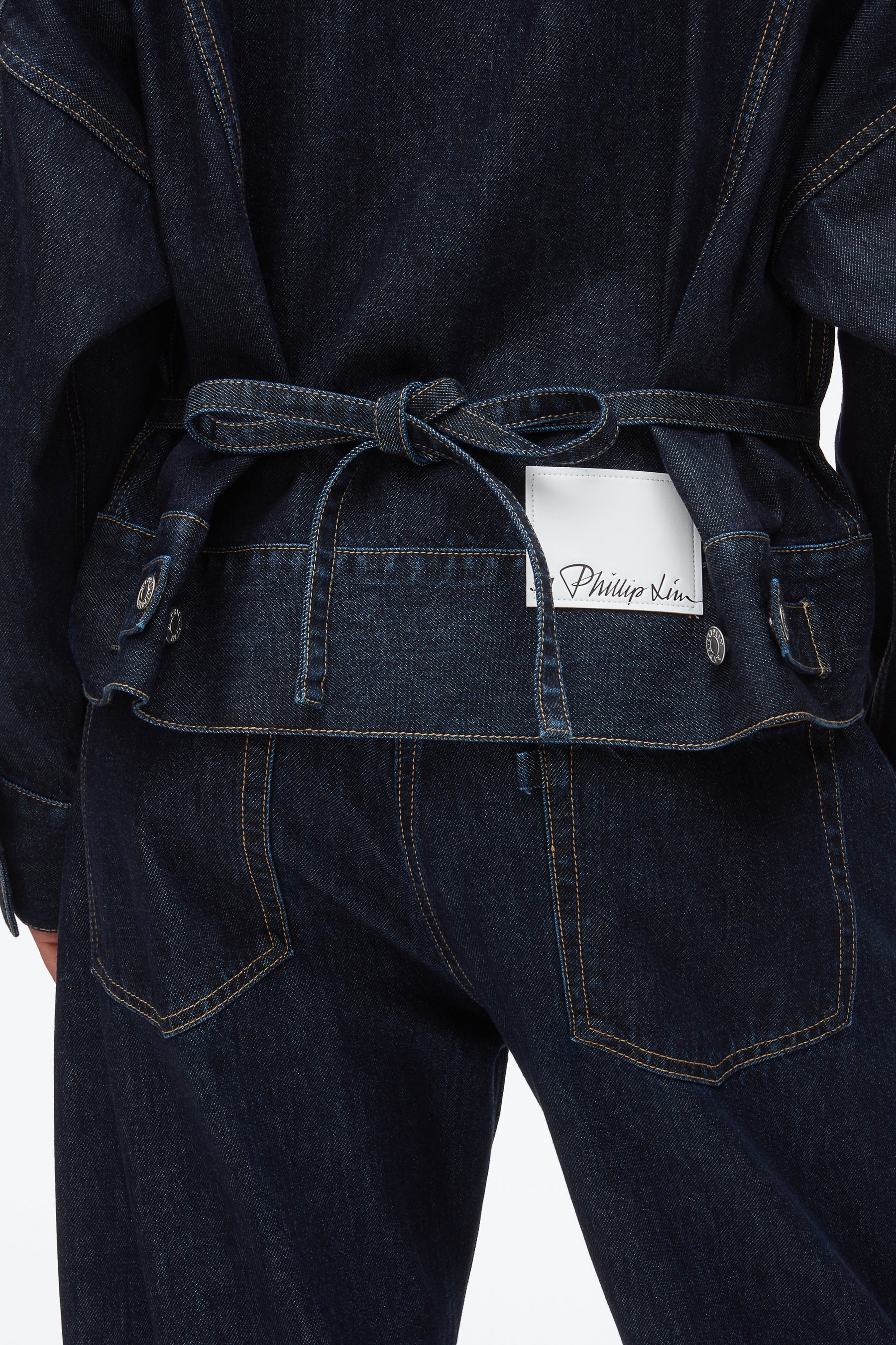 A person seen from behind wears a boxy indigo cotton denim jacket by 3.1 Phillip Lim, belted at the waist, with the label partially tucked into the waistband.