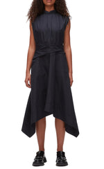 A woman in the Ruched Handkerchief Dress by 3.1 Phillip Lim, a sleeveless black midi made from technical poplin with a handkerchief hemline, paired with black loafers, stands against a plain background.