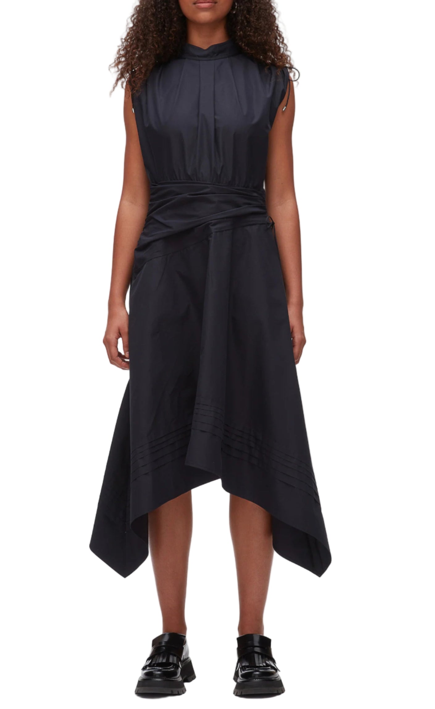 A woman in the Ruched Handkerchief Dress by 3.1 Phillip Lim, a sleeveless black midi made from technical poplin with a handkerchief hemline, paired with black loafers, stands against a plain background.