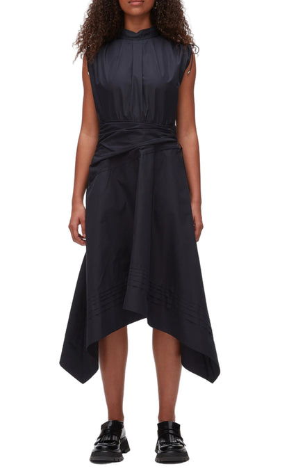 A woman in the Ruched Handkerchief Dress by 3.1 Phillip Lim, a sleeveless black midi made from technical poplin with a handkerchief hemline, paired with black loafers, stands against a plain background.