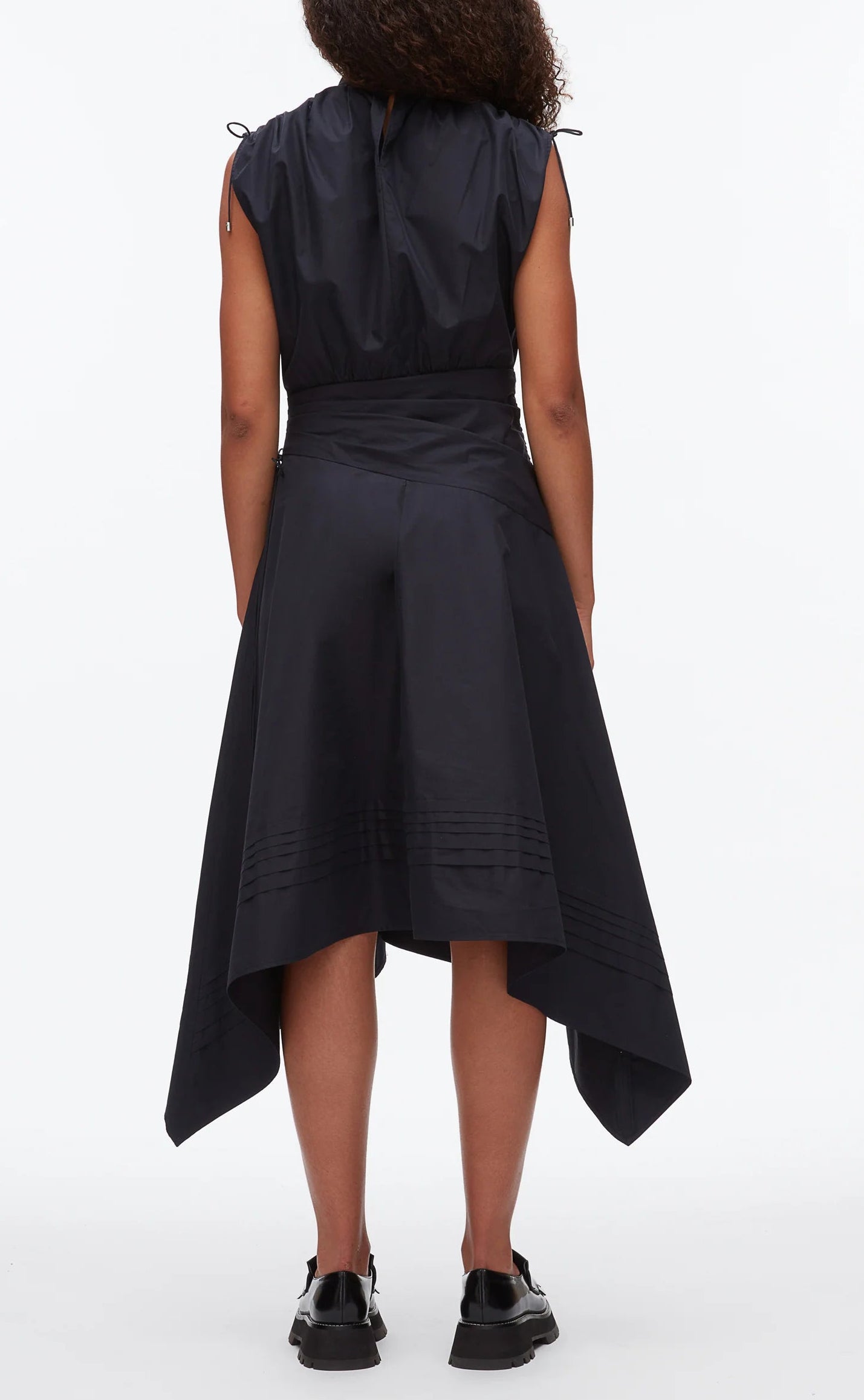A person faces away in a sleeveless black Ruched Handkerchief Dress by 3.1 Phillip Lim, crafted from technical poplin with a handkerchief hemline, paired with black shoes against a plain white background.