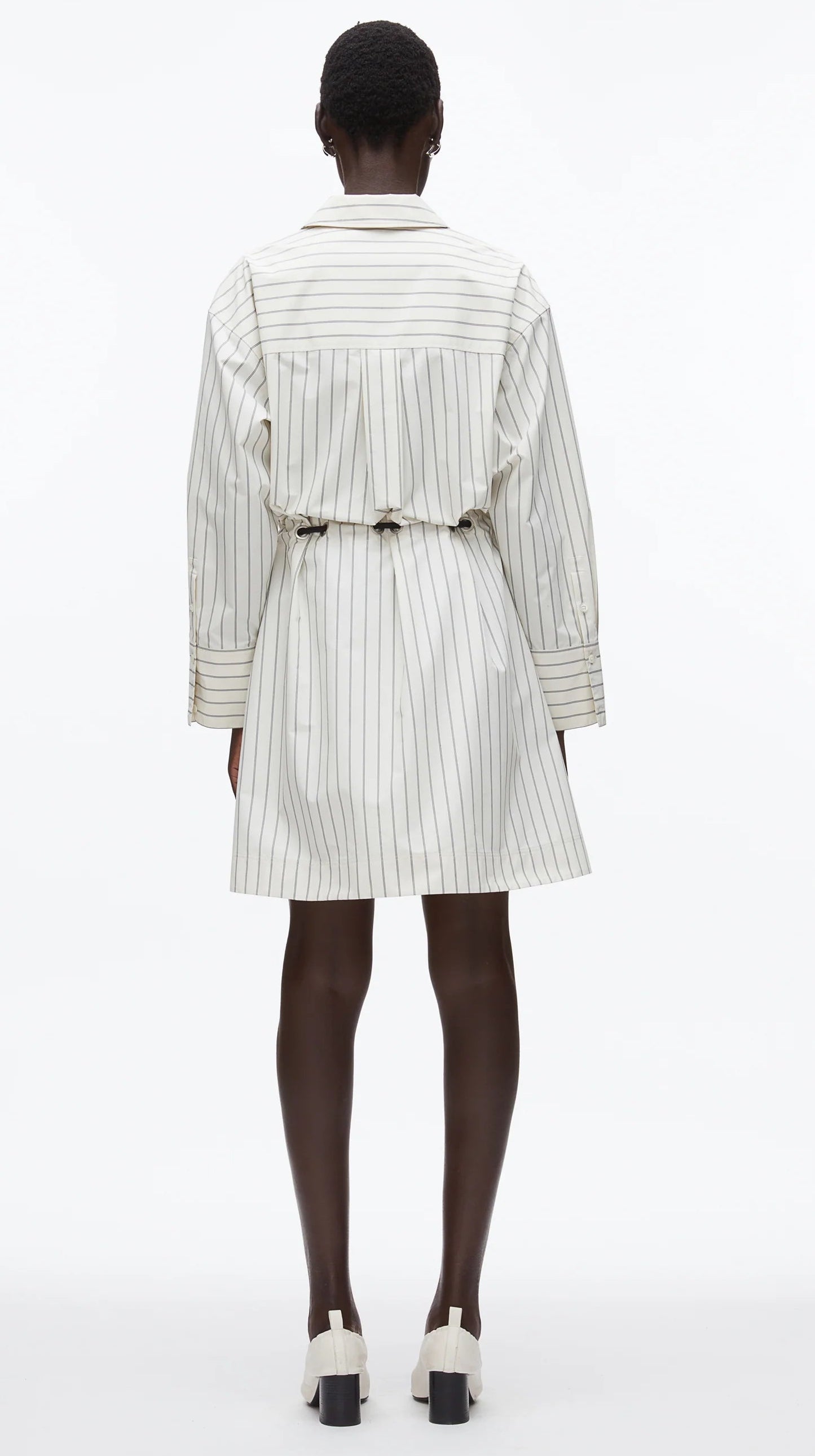 A person stands with their back to the camera, wearing the 3.1 Phillip Lim Belted Mini Shirt Dress—a long-sleeve, striped poplin dress—paired with white heeled shoes.