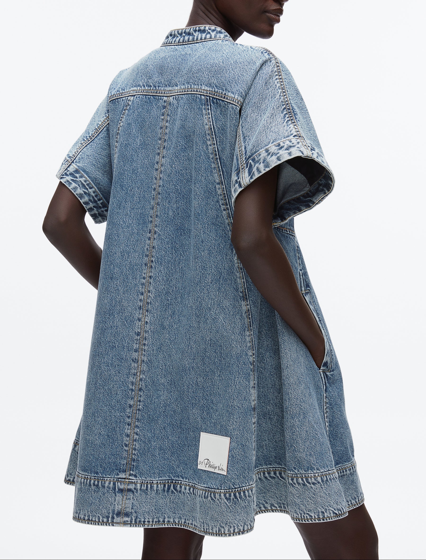 A person wears the 3.1 Phillip Lim Trapeze Light Denim Dress, featuring a relaxed trapeze silhouette, short sleeves, side pockets, and a visible white label near the hem.