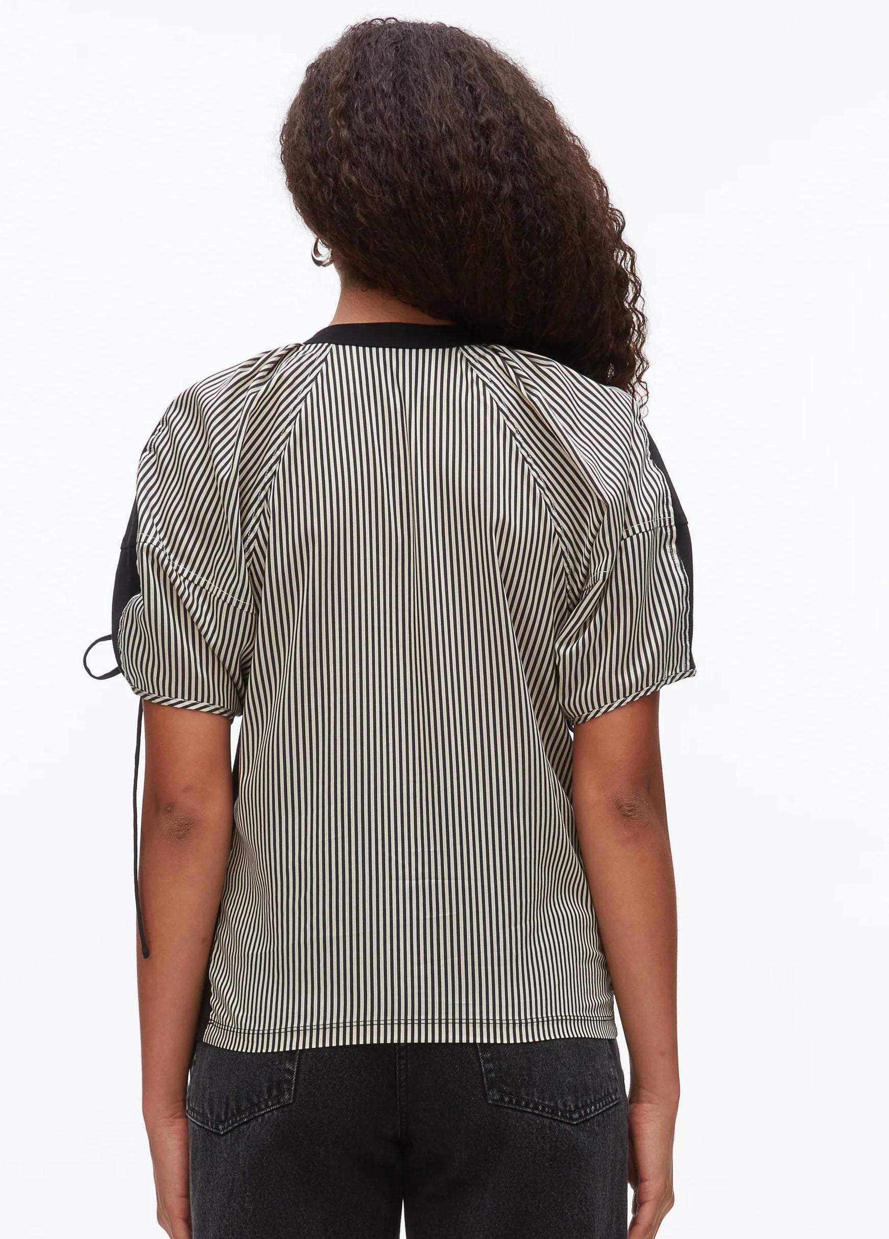 A person with long curly hair is seen from the back, wearing a 3.1 Phillip Lim Ruched T-Shirt with Combo Panel Back and dark jeans against a plain background.