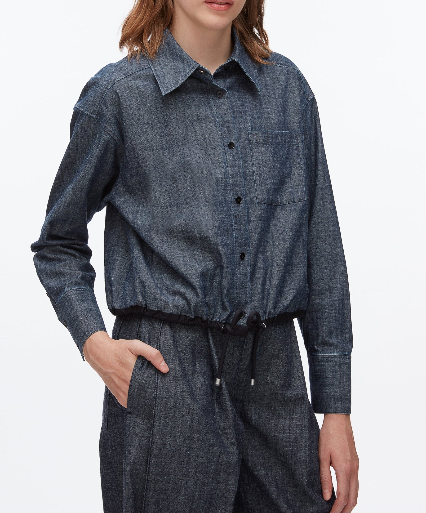 Wearing the 3.1 Phillip Lim Denim Cropped Shirt with a drawstring waist and matching pants, a person stands stylishly with one hand in their pocket.