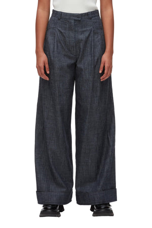 A person wears the 3.1 Phillip Lim Denim Double Pleat Wide Leg Pant in dark denim, featuring cuffed hems, paired with black shoes. The photo captures a modern yet classic silhouette from shoulders to feet against a white background.