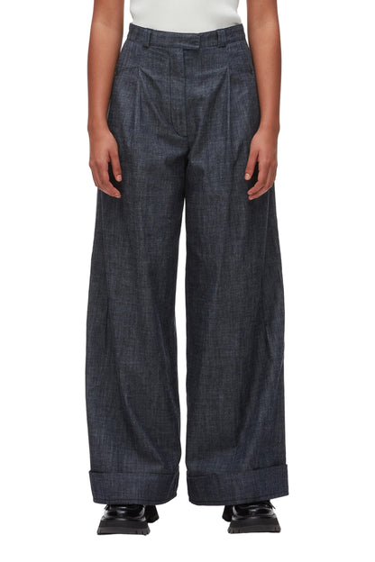 A person wears the 3.1 Phillip Lim Denim Double Pleat Wide Leg Pant in dark denim, featuring cuffed hems, paired with black shoes. The photo captures a modern yet classic silhouette from shoulders to feet against a white background.