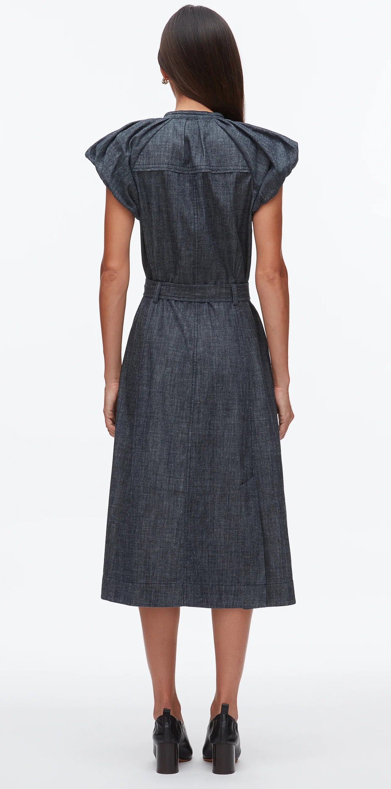 A woman faces away, wearing the 3.1 Phillip Lim Denim Puff Sleeve Dress with a romantic shoulder line, belted waist, and black heeled shoes.